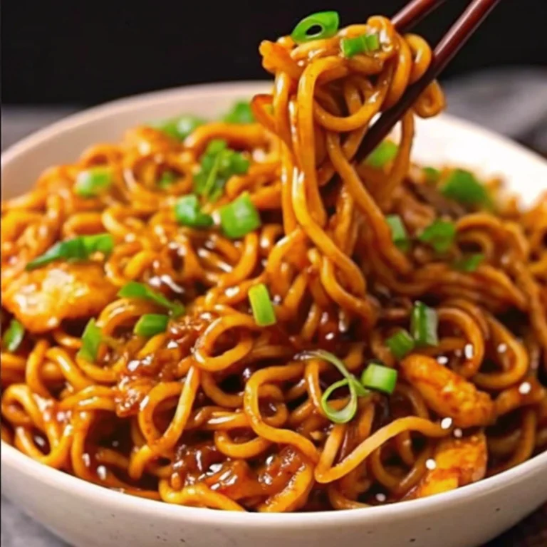 Sticky Garlic Chicken Noodles in a black skillet, showing glazed chicken pieces and noodles coated in a shiny sauce, garnished with green onions and sesame seeds.